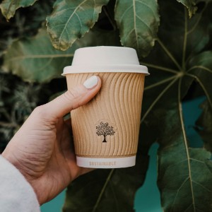 Hand hält Kaffee-Becher mit Aufdruck/Prägung "sustainable" vor grünen Pflanzen.